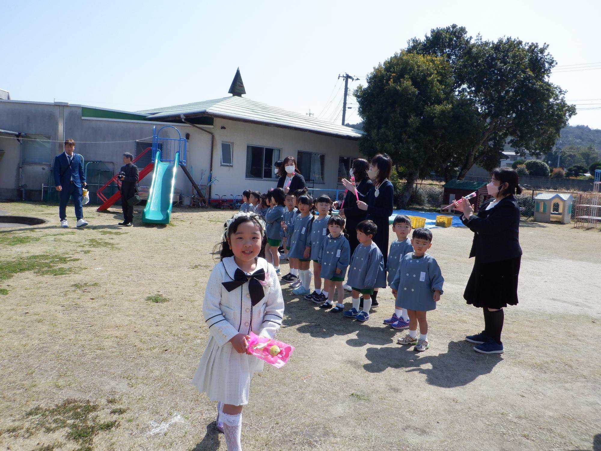 可愛いい白のツーピース姿で在園児の前をあるくおんなの子、手に花束
