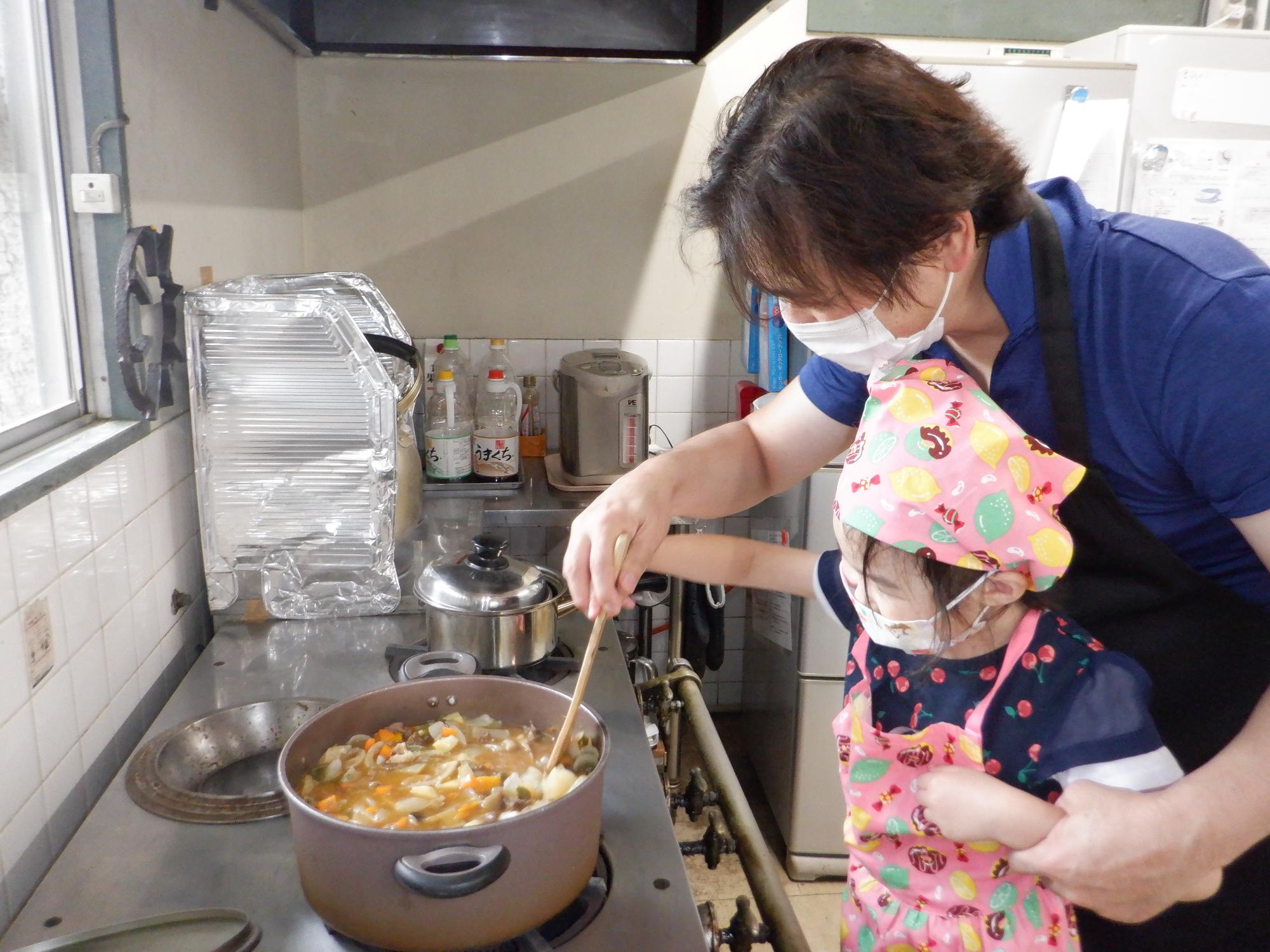 お鍋にカレールーを入れて混ぜる、先生に手を添えてもらい混ぜている女の子、