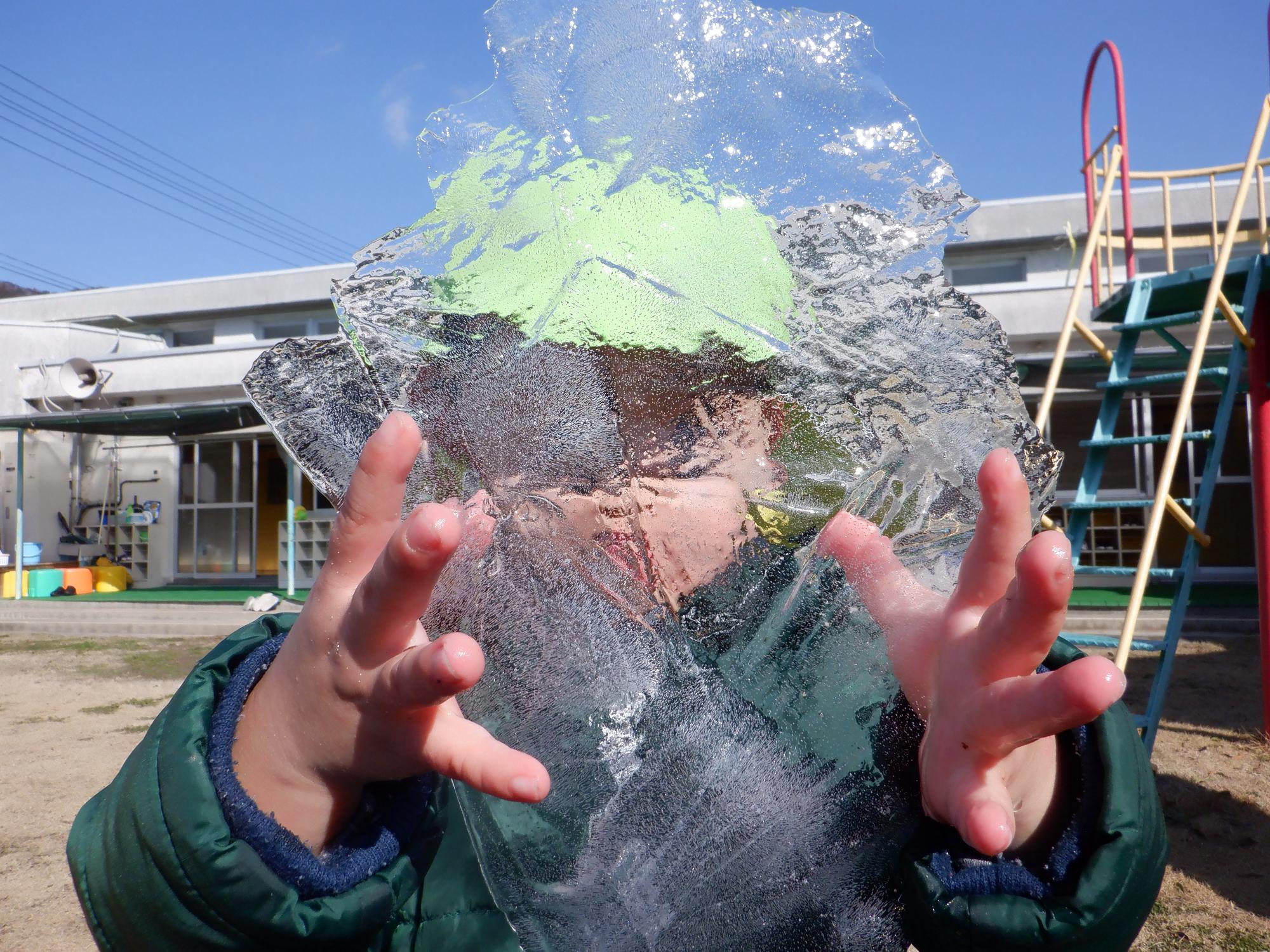 園庭で見つけた氷を顔の前でお面のようにして見せている