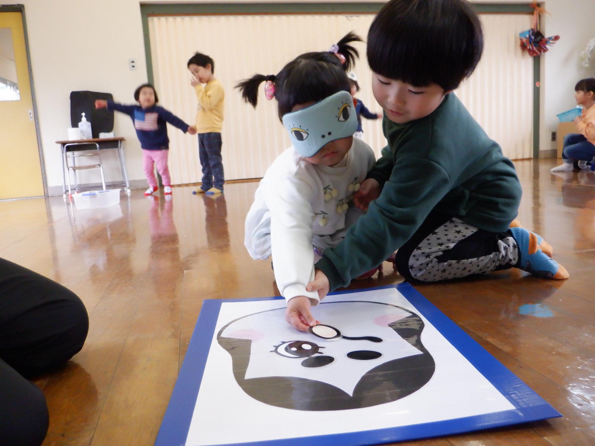 福笑い、子どものひとりが目隠し一人がガイドで鼻の位置を決めている。